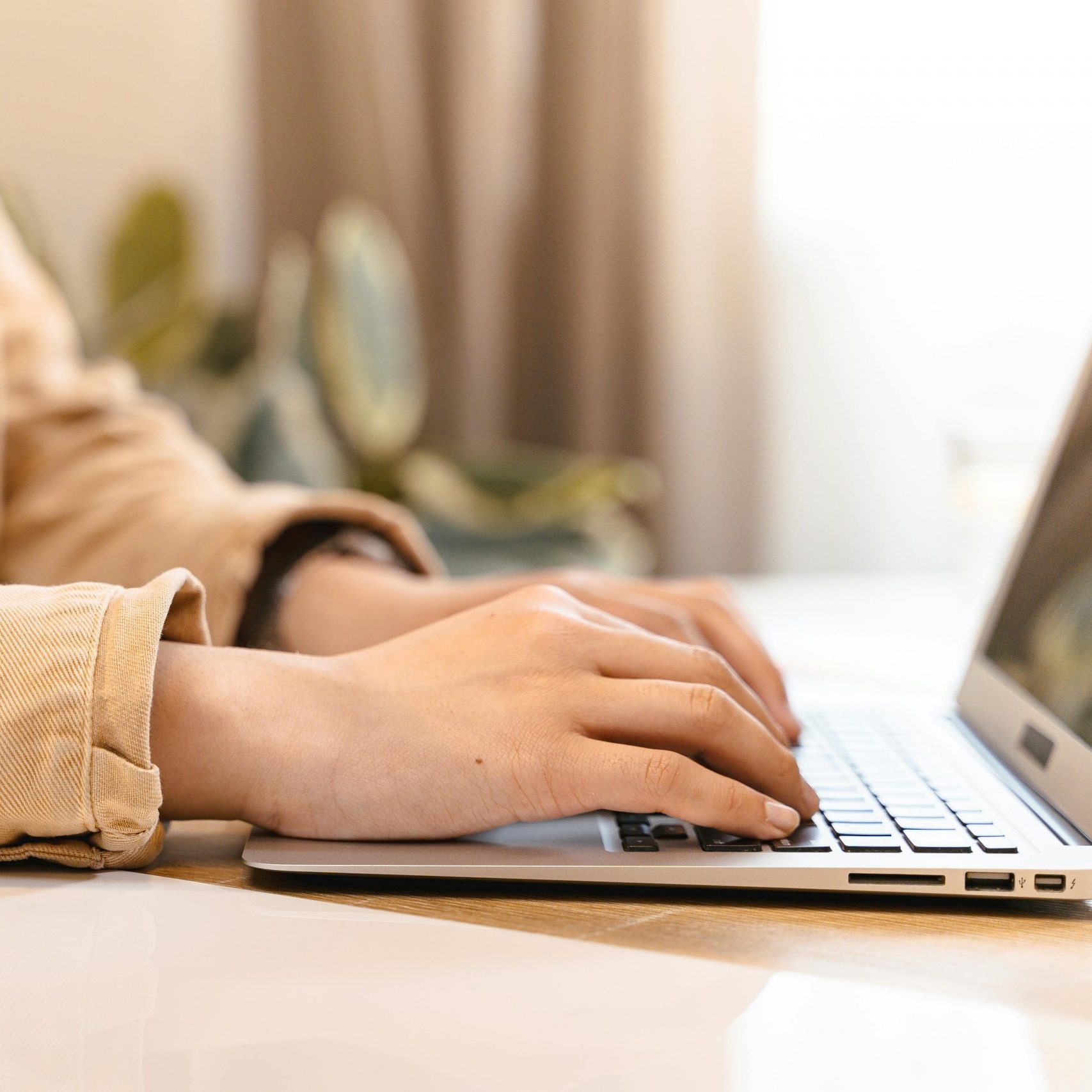 Close-up of hands typing on a laptop indoors, wearing a casual jacket.